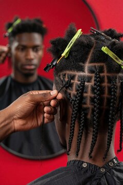 Close up back view of young afro man getting hair braids in pro salon. Hairstylist works on cornrows with hair clips. Male client sees reflection in mirror. Pro grooming, hair care service in beauty