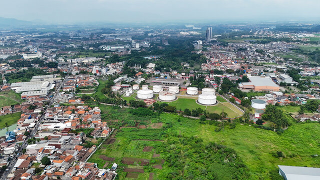 Aerial view of industrial storage tanks surrounded by residential buildings, lush green fields, and urban infrastructure in Ngamprah, West Java, Indonesia.