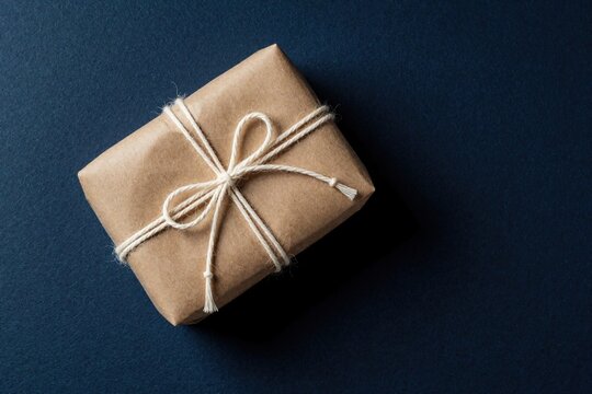 Eco friendly gift wrapped in brown kraft paper with white string twine. Minimalist present box sits on dark blue background. Sustainable package design for holiday celebration birthday surprise.
