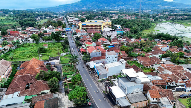 Aerial view of Tanjungsari featuring a main road, dense residential housing with terracotta rooftops, lush greenery, and distant mountains, Sumedang, West Java, Indonesia.