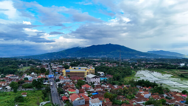 Aerial view of Tanjungsari featuring residential buildings, lush green rice paddies, and a prominent mountain under a cloudy sky, Sumedang, West Java, Indonesia.