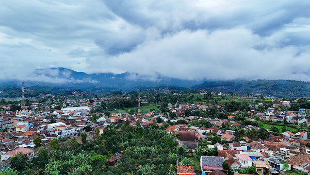 Aerial view of Tanjungsari town nestled among lush greenery and rolling hills under a dramatic cloudy sky in Sumedang, West Java, Indonesia.