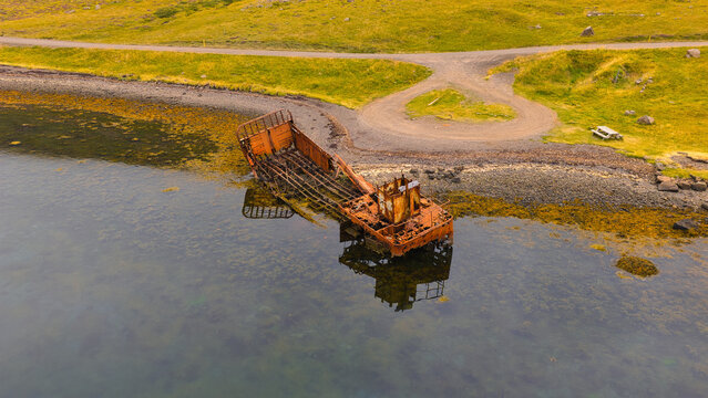 Aerial view of the rusted WWII US Navy LCM ship wreck resting in the shallow coastal waters near a grassy shoreline in Reydarfjordur, Fjardabyggd, Iceland.