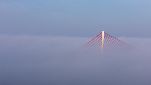 Aerial view of the cable-stayed bridge pylon and red cables emerging from a thick layer of morning fog in Gdansk, Poland.