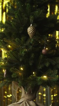 Vertical video. Woman decorating Christmas tree, hanging golden Christmas ornaments.