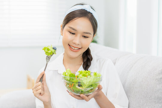 Diet, Dieting asian young woman, girl hand use fork at lettuce on mix vegetables, green salad bowl, eat food low fat to good body sitting on sofa at home. Nutritionist female, Weight loss for healthy.