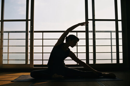 Woman doing side stretch yoga pose in silhouette before large glass windows