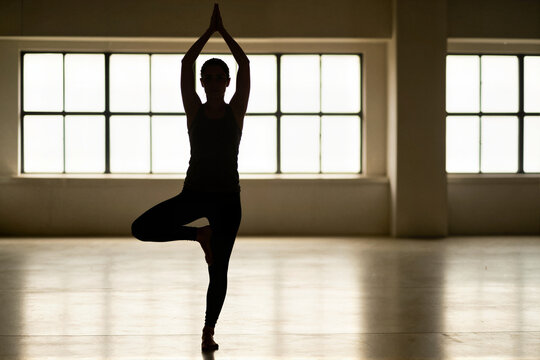 Woman in tree pose silhouette practicing yoga by large bright windows in studio