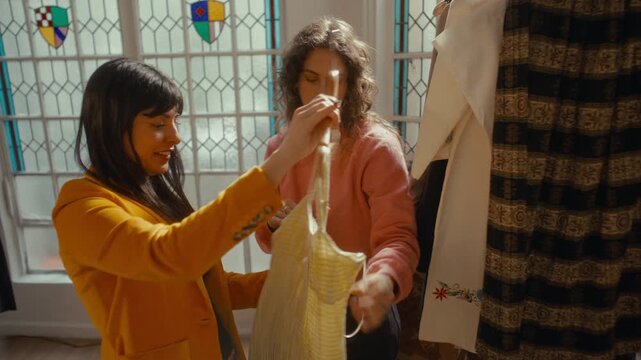Young woman holding up yellow gingham dress on hanger and discussing material and straps with female friend during shopping day at vintage thrift shop
