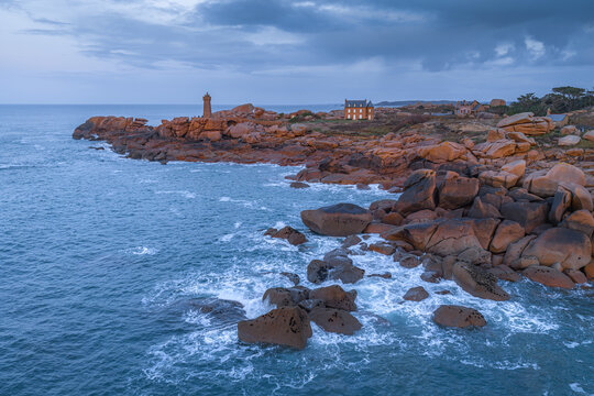 Aerial view of Phare de Mean Ruz lighthouse on the pink granite coast with rocky cliffs, crashing waves, and a historic house in Perros-Guirec, Brittany, France.
