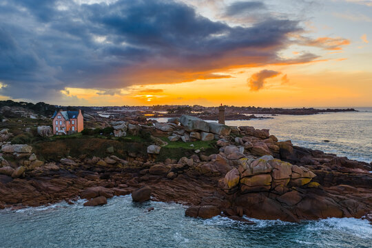 Aerial view of Phare de Mean Ruz lighthouse on the rocky coast of Perros-Guirec at sunset with dramatic clouds and granite boulders, Perros-Guirec, Brittany, France.