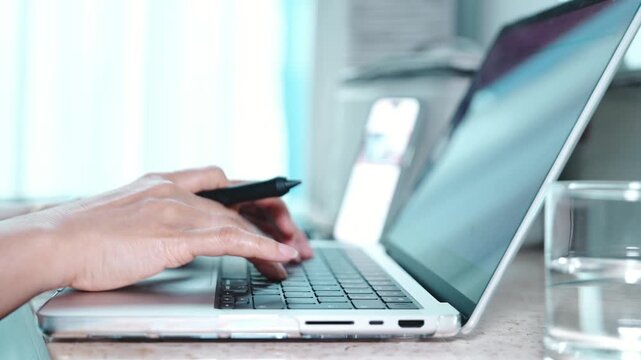 Close up of hands typing on a laptop keyboard, switching to touchpad and writing notes at a home workspace, with a glass of water nearby, showing remote work, multitasking and daily productivity.
 Cop