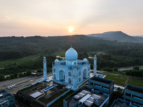 Aerial view of Budi Guna Mosque with its white dome and minarets, surrounded by lush green hills and a river at sunset in Citeureup, West Java, Indonesia.