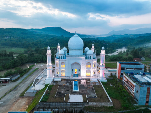 Aerial view of Budi Guna Mosque with its white dome and minarets, surrounded by lush green hills, a river, and a cloudy sky in Citeureup, West Java, Indonesia.