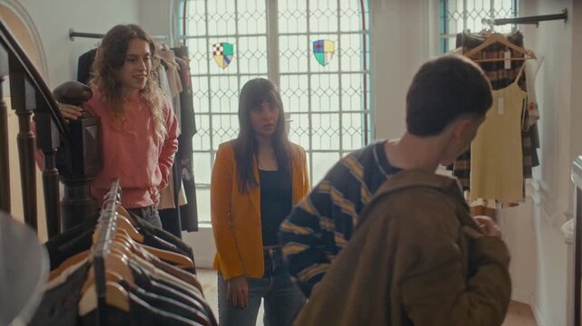 Young man trying on brown suede jacket while two female friends watching and discussing his outfit, enjoying their time shopping together in vintage thrift shop