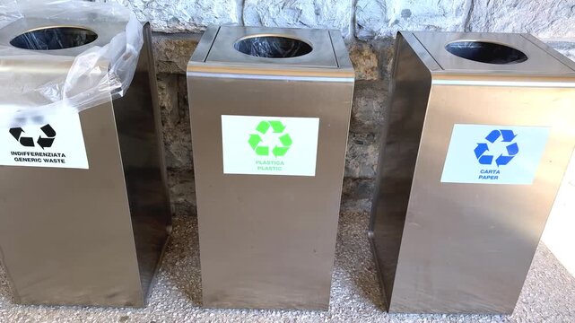 Three grey recycling bins with sorting symbols lined up outdoors near a stone wall, waste sorting and ecology concept.
