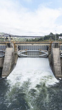 Aerial view of the Velle Dam on the Mino River with water rushing through the spillway, concrete structures, and surrounding green hills in Ourense, Ourense, Spain.