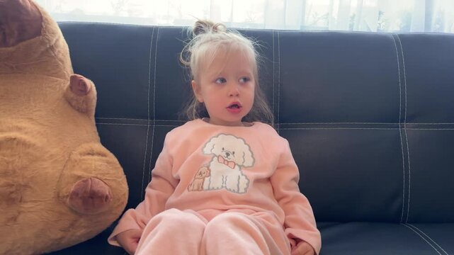 A girl sticks out her tongue and lifts her knees beside a plush capybara on a sofa, childhood humor, playful expressions, home leisure.