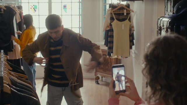 Young woman recording smartphone video of her male friend trying on brown jacket, capturing social media content for vlog about vintage thrift shop