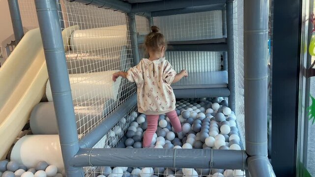 A girl walks through a ball pit and climbs padded steps beside a slide, indoor playground, active childhood, motor development.