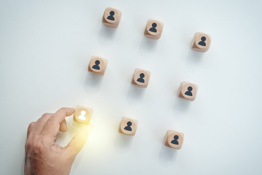 Hand selecting a white highlighted human icon wooden block among black icon blocks on a white desk, concept of human resource management, talent acquisition, and recruitment.
