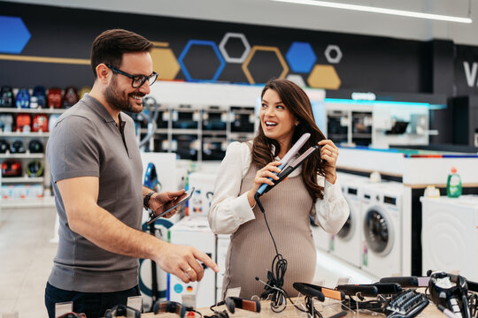 Helpful male clerk advising a happy expectant mother holding a styling iron while shopping for home appliances in a brightly lit electronics marketplace.