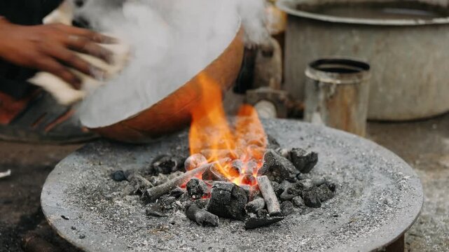 Indian craftsman tinning a copper bowl over a traditional charcoal fire in a workshop, creating handmade metal cookware