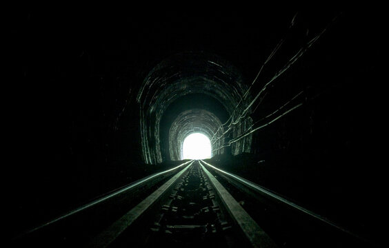 Railway tunnel in Thailand built in 1914, with steel tracks and sleepers extending through a dark arched interior toward a bright exit, alongside cables running along the weathered tunnel wall.
