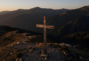 Fototapeta premium Christianity and faith concept, wooden cross on a mountain peak at sunset with winding road and vast landscape for spiritual journey and hope