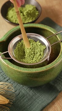 Pressing and sifting bright green matcha powder through metal mesh sieve into green ceramic bowl chawan with bamboo wooden spatula scoop chashaku. Traditional Japanese matcha tea preparation.