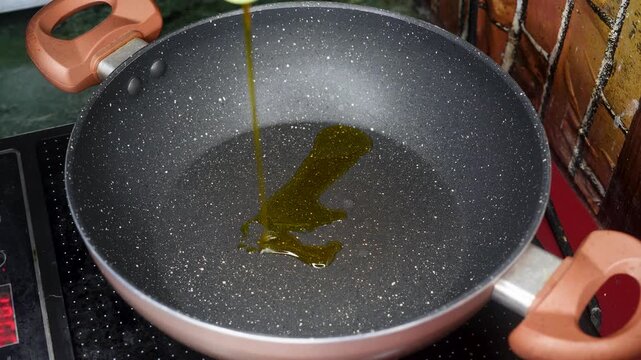 Pouring golden cooking oil, a bottle of oil is being poured into a clean nonstick pan on a induction oven, indicating a preparation for cooking at home kitchen.