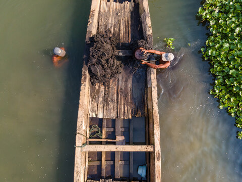 Aerial view of Kalirajut with two men dredging mud from a wooden boat in the river, surrounded by water hyacinths, Patikraja, Central Java, Indonesia.