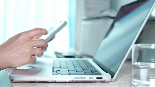 Close up of hands using a smartphone and laptop, typing messages on a mobile phone, working with a keyboard and touchpad, and pointing at the screen in a modern home office workspace.