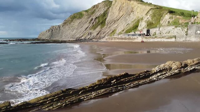 Low Drone Flight over Zumaia Beach with Wet Sand Reflection and Flysch Cliffs, Basque Country. Ideal for atmospheric intros, travel vlogs, coastal tourism ads and nature films.