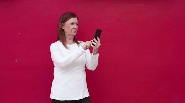 Mature woman texting on smartphone, isolated against a red wall
