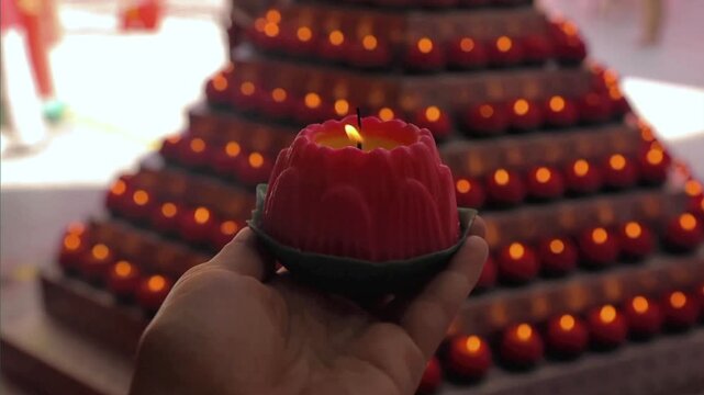 A hand holding a glowing red lotus candle, with rows of blurred, warm lights in the background suggesting a temple or spiritual setting