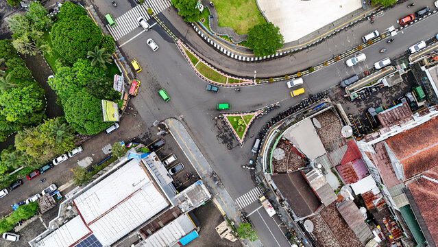 Aerial view of a busy street intersection featuring vehicles, pedestrian crossings, surrounding buildings with tiled roofs, and lush green trees in Sukabumi, West Java, Indonesia.