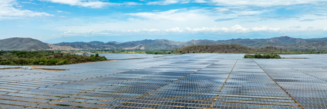 Aerial view of a large scale photovoltaic power plant with rows of solar panels extending across a landscape with rolling hills under a blue sky.