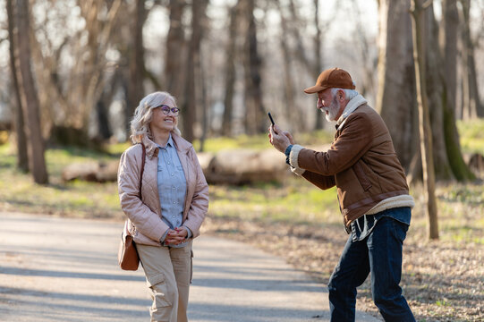 Happy senior man taking a smartphone photo of a smiling mature woman posing on a park path during a sunny day, enjoying photography and leisure time together