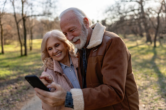 Happy senior couple smiling and looking at a smartphone together while embracing in a sunny park, enjoying technology and sharing a joyful moment outdoors