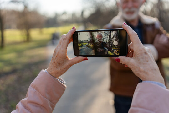 Happy senior man with white beard smiling and posing for a smartphone photo taken by a woman outdoors in a sunny park, enjoying a relaxed moment together