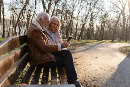Happy senior couple using a smartphone together on a park bench, illustrating online banking, mobile financial services and digital independence during retirement