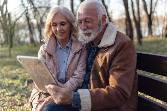 Smiling senior man and woman spending quality time together outdoors, sharing a tablet while relaxing on a park bench in warm natural sunlight