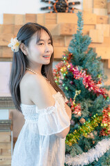 Profile portrait of a beautiful young Asian woman wearing a white off-the-shoulder top and a white flower in her hair, smiling while standing beside a decorated Christmas tree in an indoor setting © maemodnit13