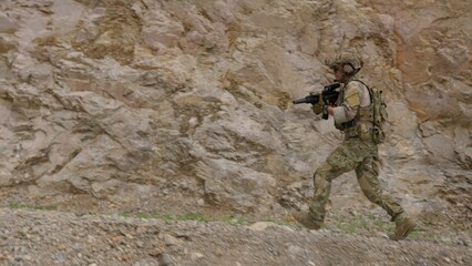 Fototapeta premium A special forces soldier in full combat gear advances with a rifle during a tactical mission, moving swiftly across rugged, rocky terrain with alertness and focus.