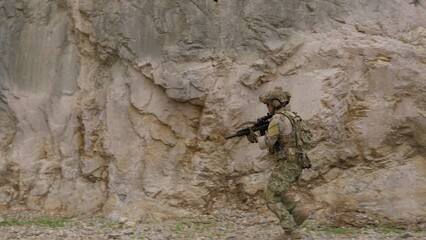 Fototapeta premium A special forces soldier in full combat gear advances with a rifle during a tactical mission, moving swiftly across rugged, rocky terrain with alertness and focus.
