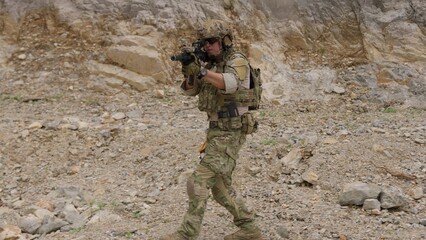 Fototapeta premium A special forces operator in full combat gear advances cautiously through a rocky, hostile environment while aiming his assault rifle on a tactical security patrol.