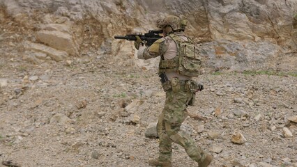 Fototapeta premium A special forces operator in full combat gear advances cautiously through a rocky, hostile environment while aiming his assault rifle on a tactical security patrol.