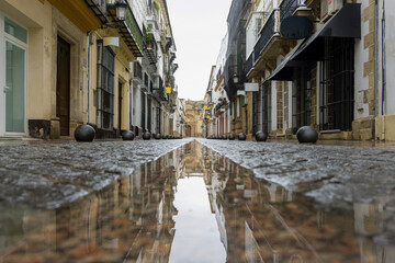 Wet cobblestone street in El Puerto de Santa Maria presenting a symmetrical reflection of traditional Spanish architecture and historic buildings under an overcast sky © photoopus