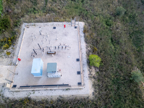 Aerial view of a fenced utility facility with industrial equipment, pipes, and control cabinets on a gravel surface surrounded by wild vegetation in Teulada, Valencian Community, Spain.
